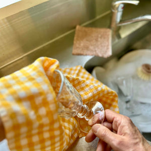 Person drying a glass with a yellow checked tea towel in a kitchen sink.