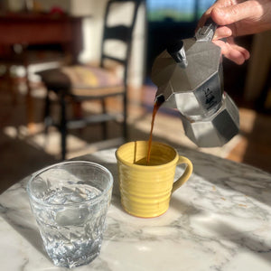 Coffee pouring from a Moka pot into a Dijon yellow mug on a marble surface.