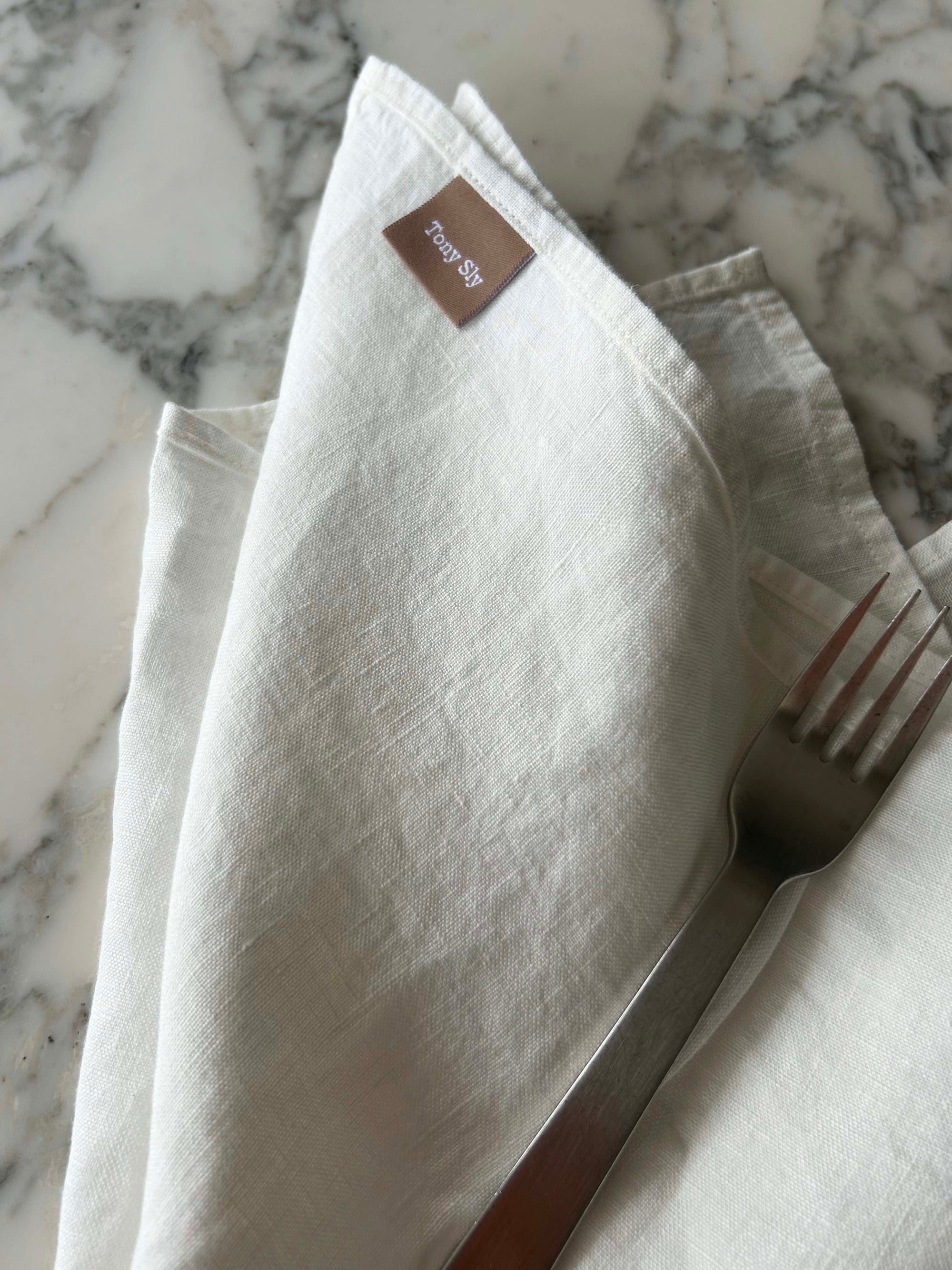 Folded white napkin with a fork on a marble surface, featuring a brand label.