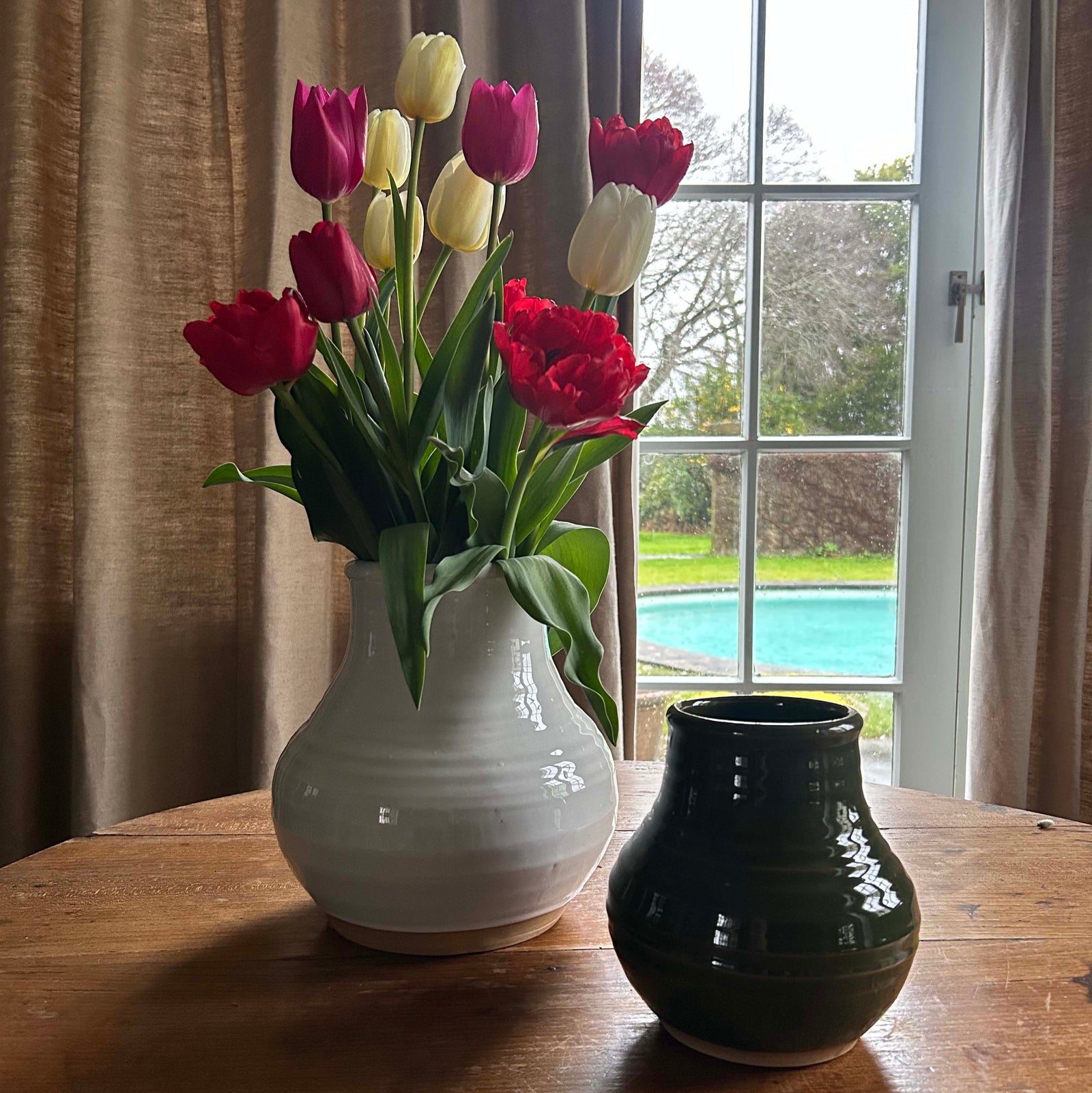White vase with red and yellow tulips on a wooden table near a window with curtains.