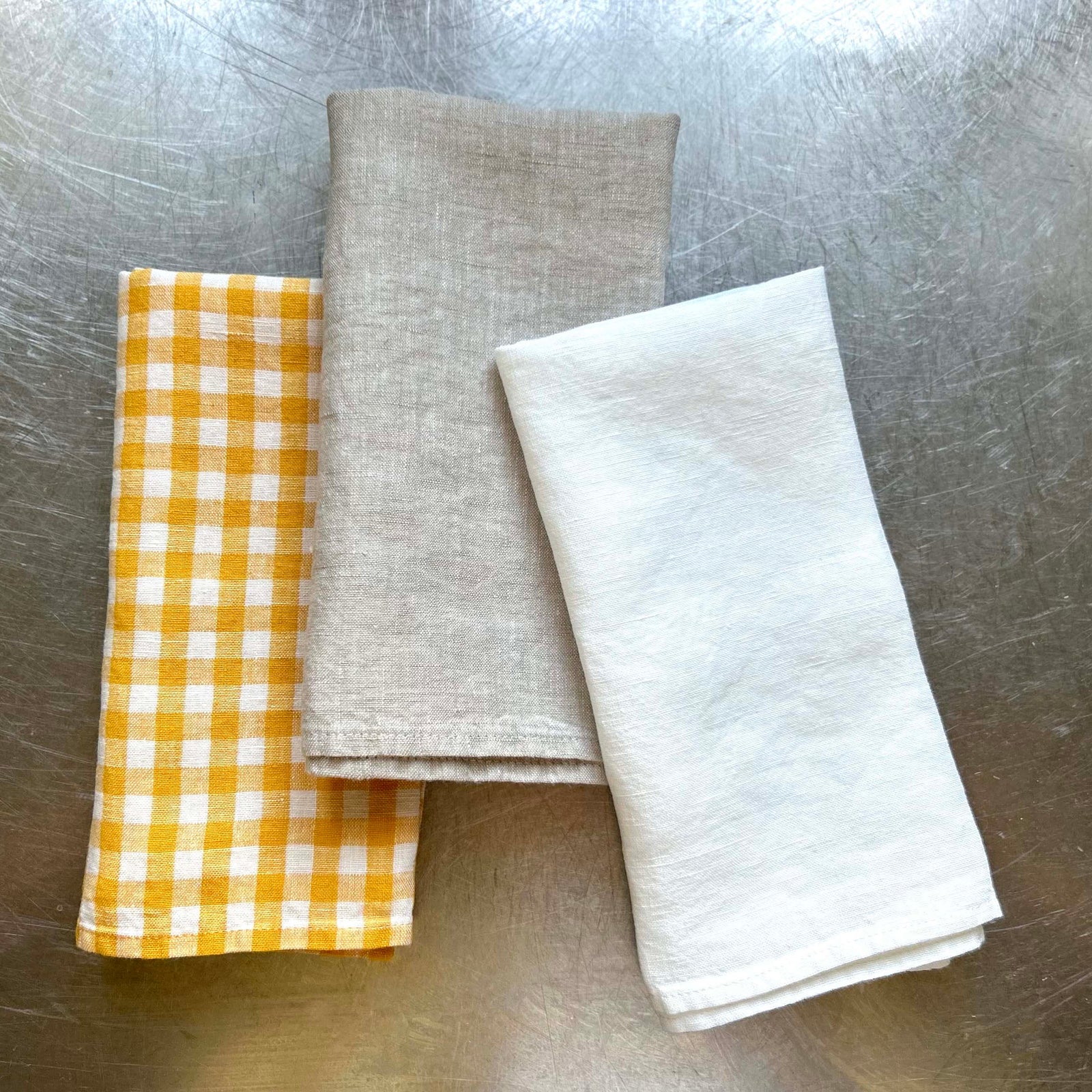 Three folded napkins in yellow check, natural, and white on a metallic surface.